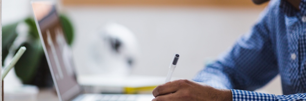 Henkilö istuu pöydän ääressä kannettavan tietokoneen edessä ja kirjoittaa kynällä muistikirjaan.