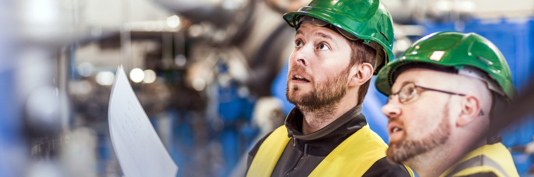 Kaksi vihreäkypäräistä työntekijää katsoo vasemmalle ylöspäin teollisuusympäristössä.