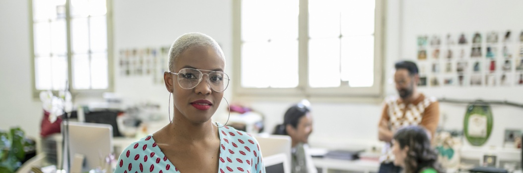 Silmälasipäinen nainen istuu etualalla pöydän ääressä ja katsoo kameraan. Taustalla kolme henkilöä juttelee keskenään toimistossa.