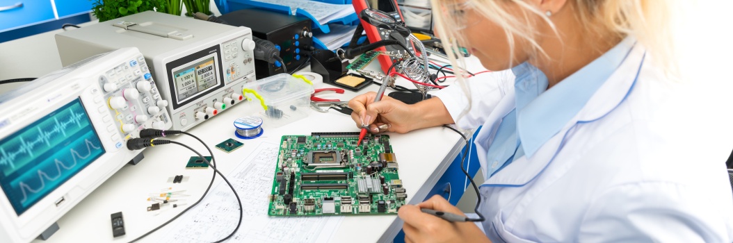 Vaaleahiuksinen henkilö istuu pöydän ääressä juottamassa piirilevyä.