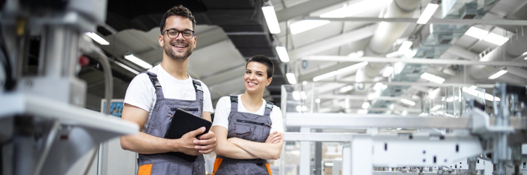 Kaksi harmaahaalarista työntekijää seisoo tuotantokoneiden edessä ja hymyilee kameralle.