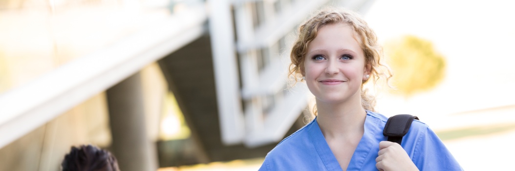 Vaaleatukkainen nainen hymyilee kameran suuntaan ja kävelee olkalaukku ja muistikirja kädessä hoitajan vaatteet päällään.