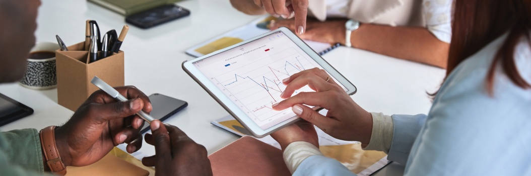 Lähikuvassa kolme henkilöä istuu pöydän ääressä ja katselee tablettilaitteella näkyvää viivakaaviota.