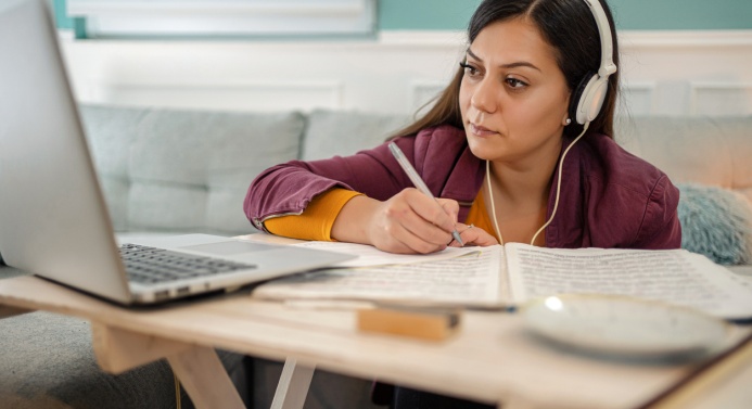 Nainen istuu kuulokkeet päässä tietokoneen ruudun äärellä ja kirjoittaa muistiinpanoja.