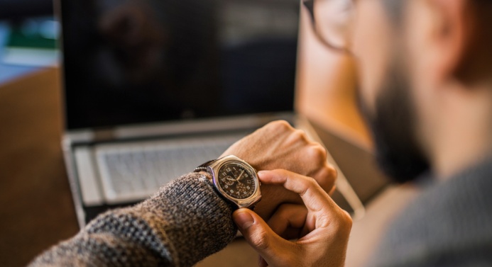 Parrakas, silmälasipäinen henkilö istuu kannettavan tietokoneen edessä ja katsoo rannekelloaan.