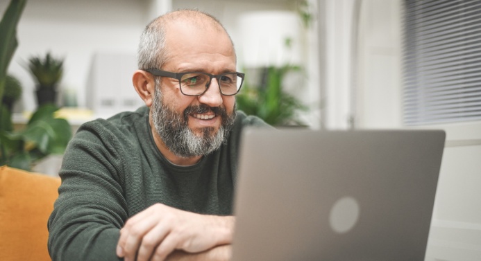 Silmälasipäinen parrakas mies istuu kannettavan tietokoneen ääressä ja hymyilee.
