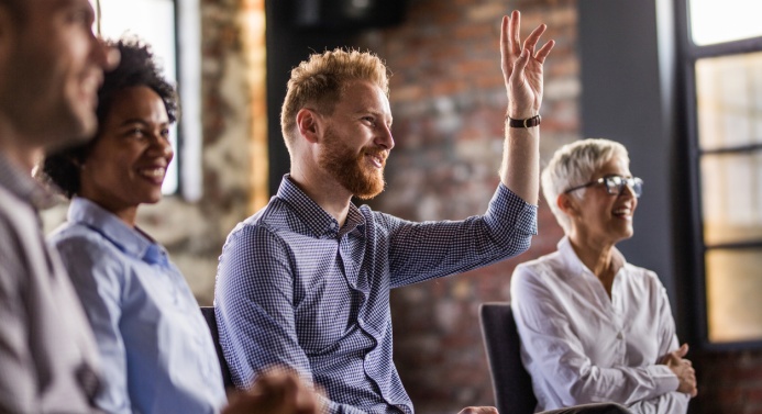 Neljä henkilöä istuu hymyillen toimistossa ja katsovat samaan suuntaan. Kuva tarkentaa yhteen henkilöön, joka viittaa.
