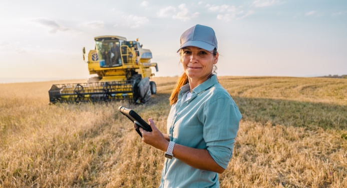 Viljapellolla seisova työntekijä katsoo kameraan älylaite kädessään. Taustalla puimuri aurinkoisessa peltomaisemassa.