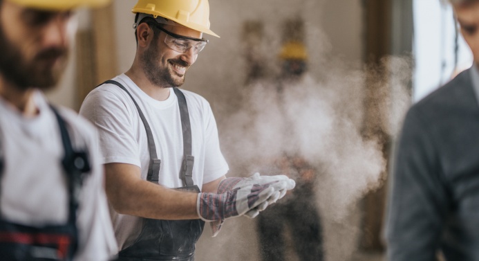 Kolme miestä rakennuskypärät ja -varusteet päällä. Kuva tarkentuu mieheen, joka hymyilee ja lyö yhteen käsiä, joista pöllähtää rakennuspölyä ilmaan.