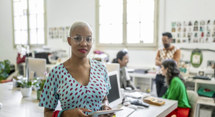 Silmälasipäinen nainen istuu etualalla pöydän ääressä ja katsoo kameraan. Taustalla kolme henkilöä juttelee keskenään toimistossa.