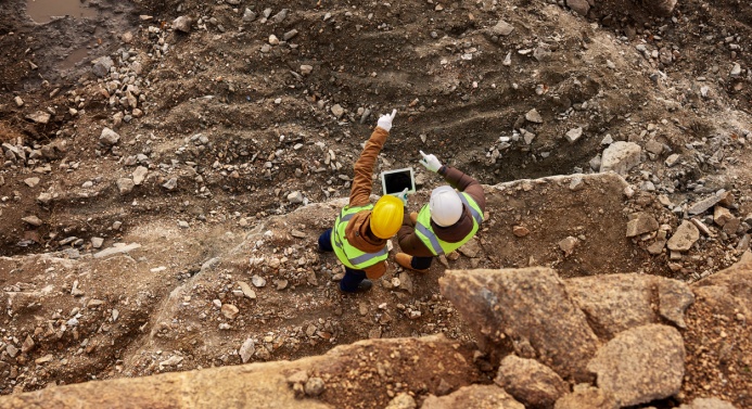 Kaksi kypäräpäistä ihmistä on kuvattu ylhäältä päin työmaalla keskustelemassa ja katsomassa tablettilaitetta. Toinen henkilöistä osoittaa sormella eteensä.