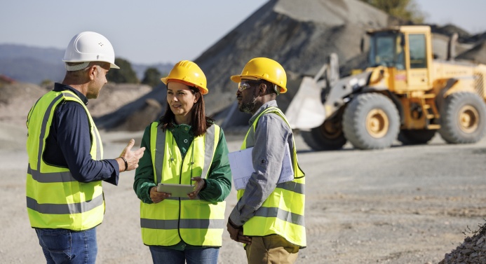 Kolme kaivostyömaalla seisovaa henkilöä keskustelee yhdessä. Taustalla kauhakuormain.