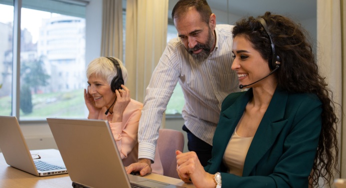 Toimiston pöydän ääressä istuu kaksi henkilöä kannettavien tietokoneiden ääressä ja yksi henkilö seisoo ja opastaa heitä.