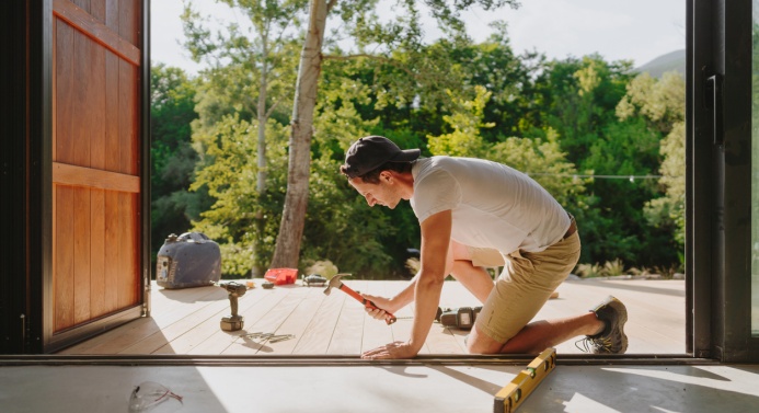 Mies rakentaa terassia ja lyö vasaralla nauloja auringonpaisteessa. Taustalla vehreä metsämaisema.
