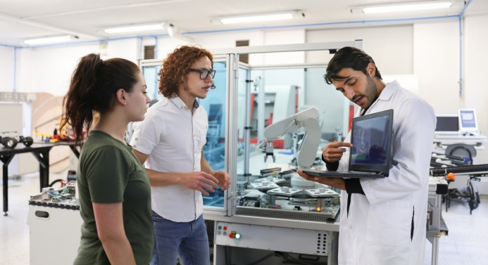 Kolme henkilöä seisoo robotin vieressä. Yksi heistä näyttää muille tietokoneen näytöltä 3D-kuvaa robotista.