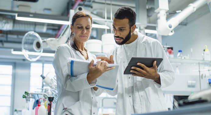 Kaksi valkotakkista henkilöä seisoo vierekkäin laboratoriossa ja katsoo lehtiötä.