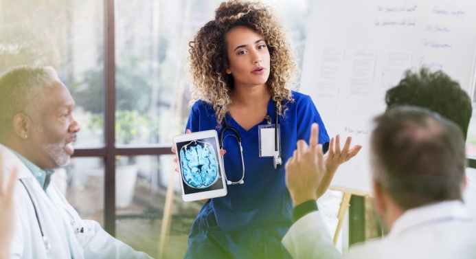 Siniasuinen nainen seisoo kokoushuoneessa valkotakkisten henkilöiden edessä ja näyttää tablettitietokoneelta kuvaa aivoista.