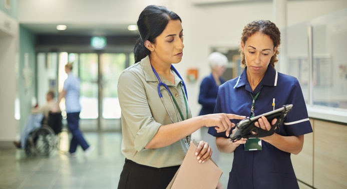 Kaksi lääketieteen ammattilaista katsoo yhdessä tablettilaitetta ja keskustelee. Toisella on kaulassaan stetoskooppi. Toisella on yllään hoitajan asu.