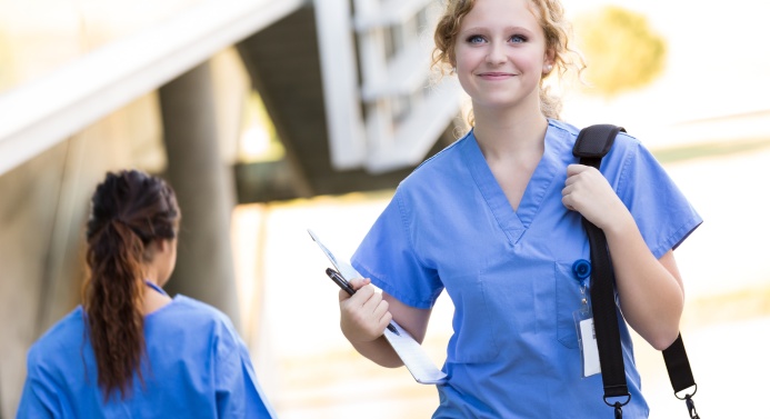 Vaaleatukkainen nainen hymyilee kameran suuntaan ja kävelee olkalaukku ja muistikirja kädessä hoitajan vaatteet päällään.