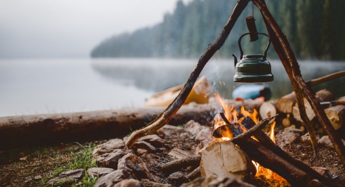 Lähikuva nuotiosta sumuisen metsämaiseman ja vesistön edustalla. Nuotion yllä roikkuu pieni kahvipannu.