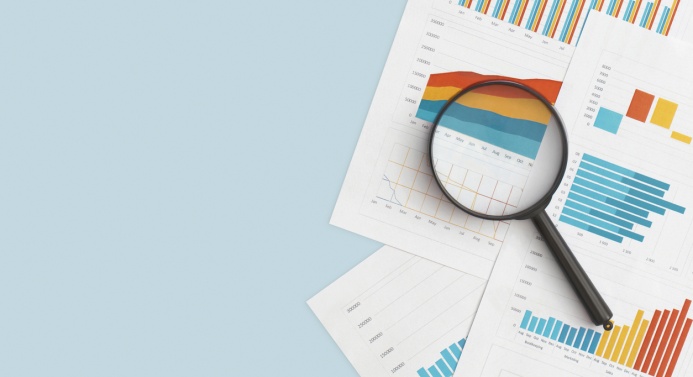 Kuvassa pöydällä papereita, joissa erilaisia kuvaajia, ja päällä suurennuslasi