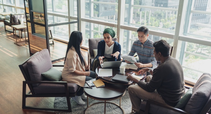 Neljä ihmistä istuu nojatuoleilla pöydän ääressä toimistossa ja keskustelee paperit edessään.