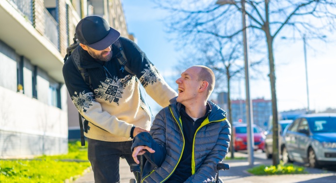 Mies työntää toista miestä pyörätuolissa aurinkoisessa säässä. Miehet hymyilevät toisilleen.